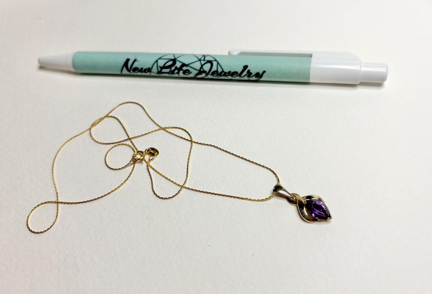 A gold necklace with an amethyst pendant and a pen with "New Life Jewelry" logo.