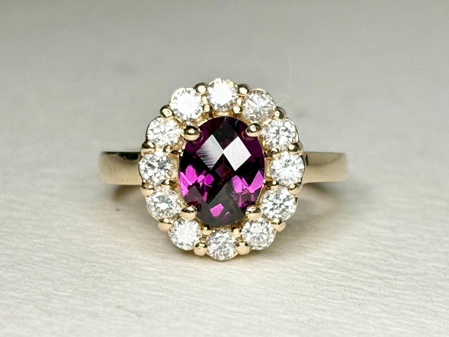 Oval checkerboard faceted rhodolite garnet ring surrounded by diamonds in a yellow gold halo setting.