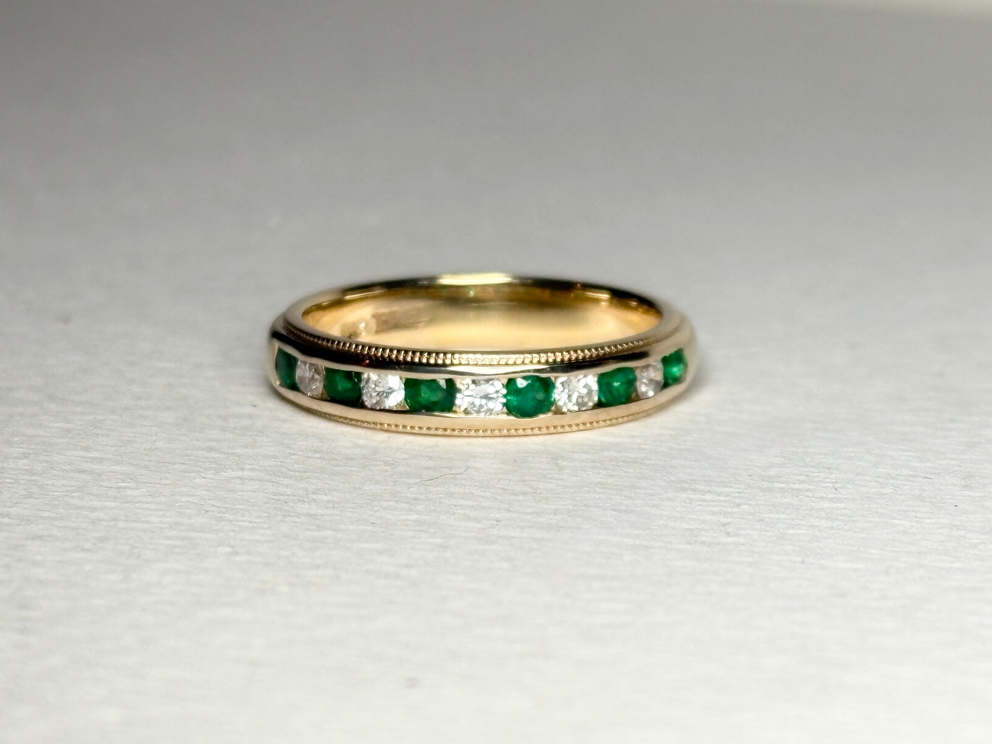 18k gold band with alternating emeralds and diamonds, featuring milgrain detailing.