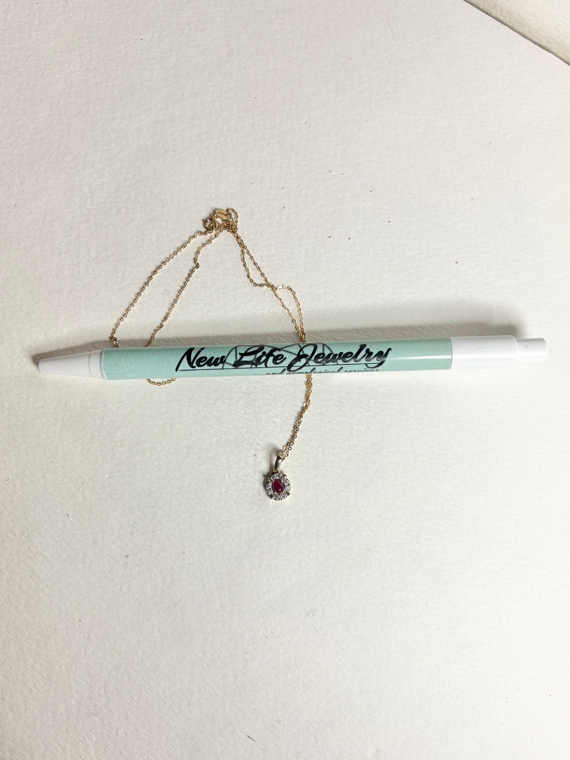 A gold necklace with a ruby and diamond pendant rests on a pen.
