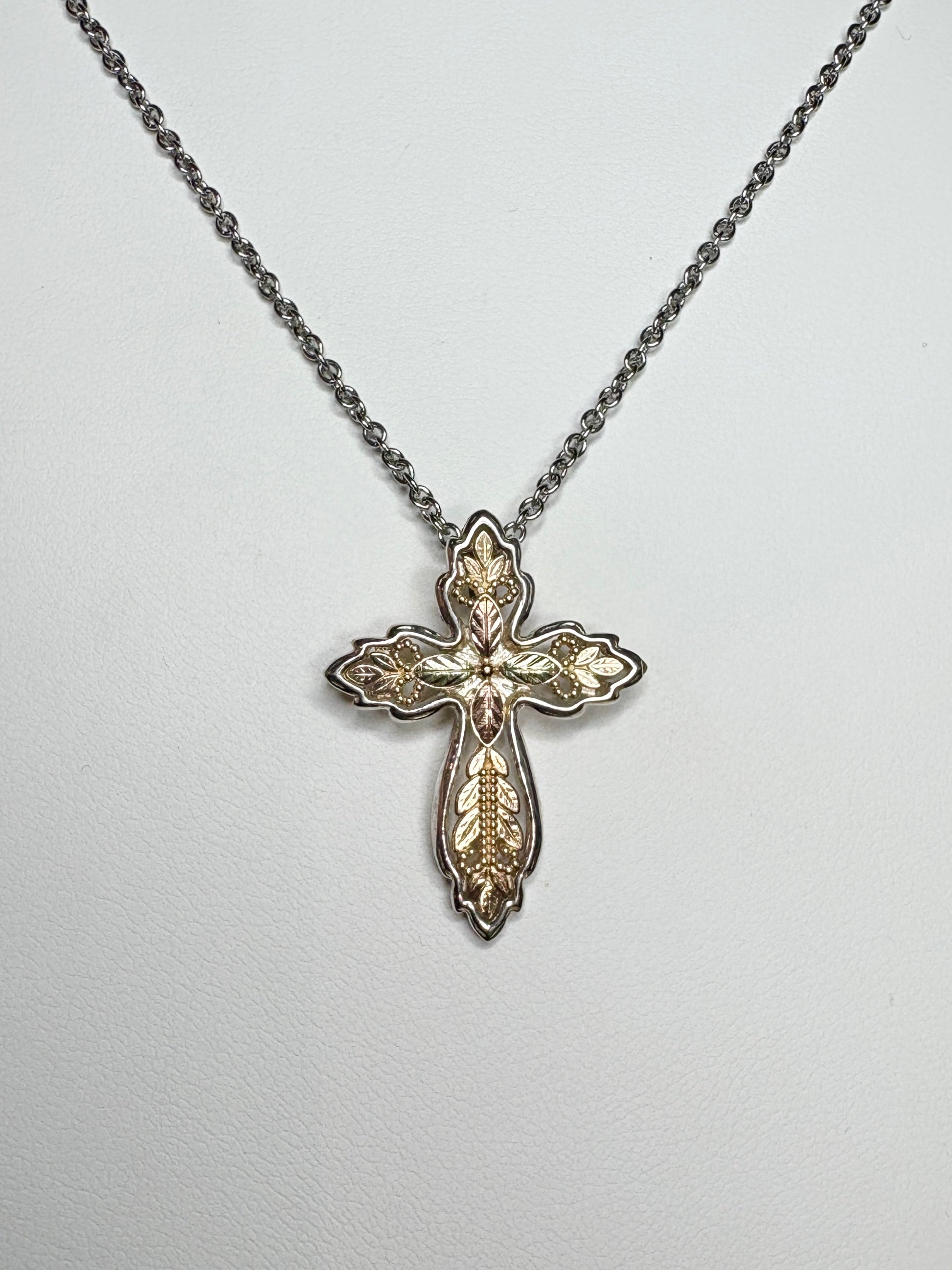 Black Hills Gold ornate cross pendant with green and rose gold leaves on a yellow gold base with a sterling silver frame.