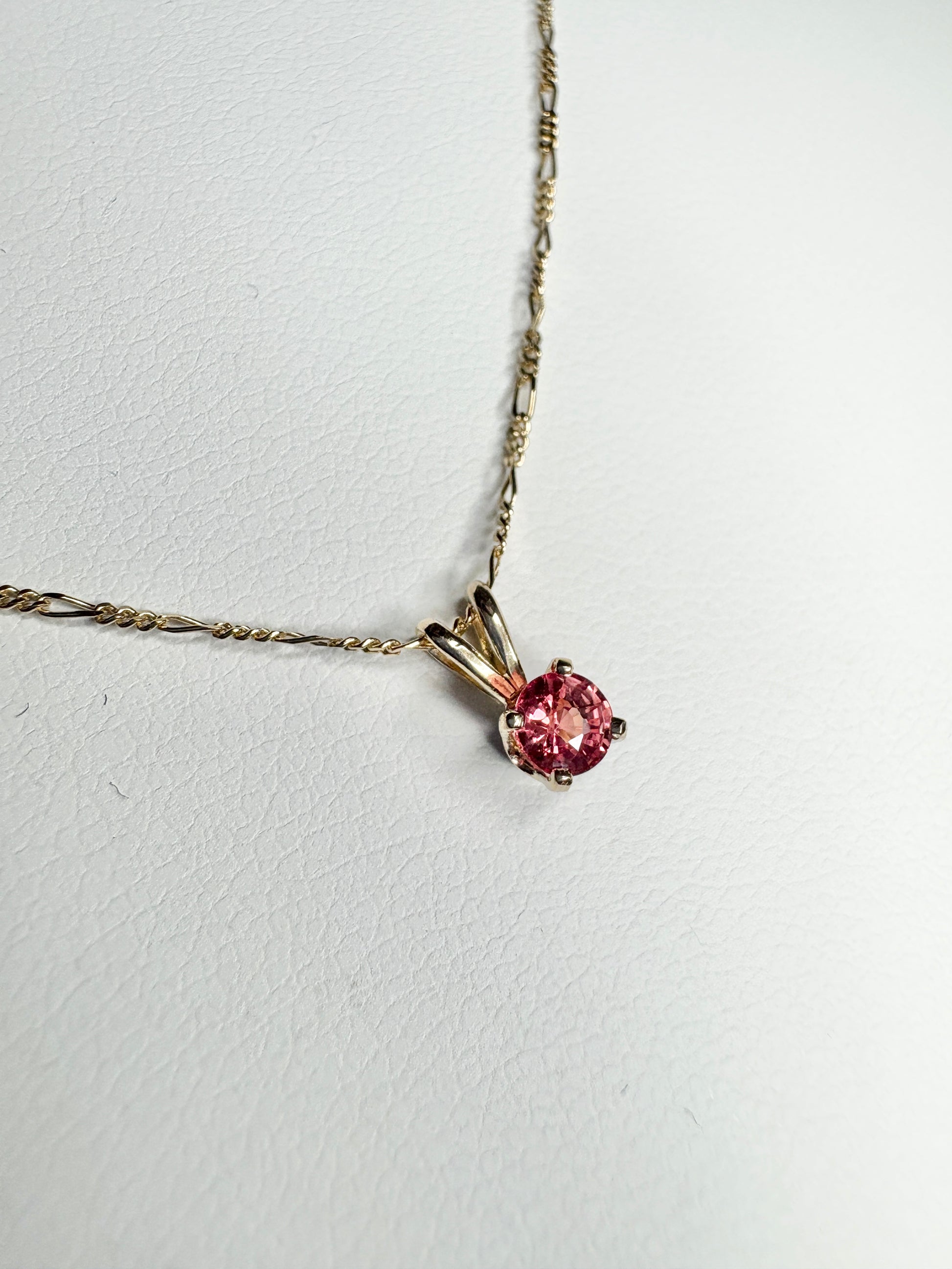 A small, round padparadcha sapphire pendant hangs from a delicate 14k gold figaro chain.