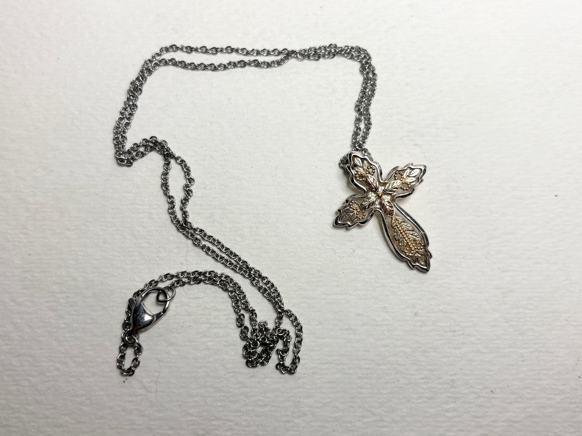 A Black Hills Gold ornate cross pendant with a sterling silver frame on a stainless steel chain.