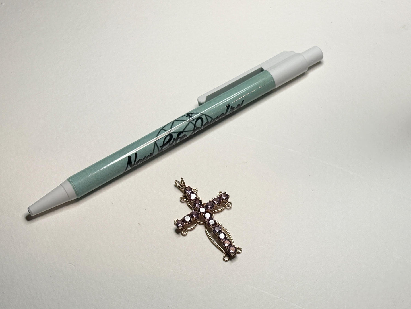 A gold cross pendant with purple stones rests on a white surface next to a pen.