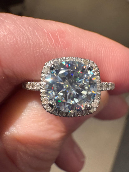 A cushion-shaped halo ring with a large moissanite center stone and small diamonds on a white gold band.