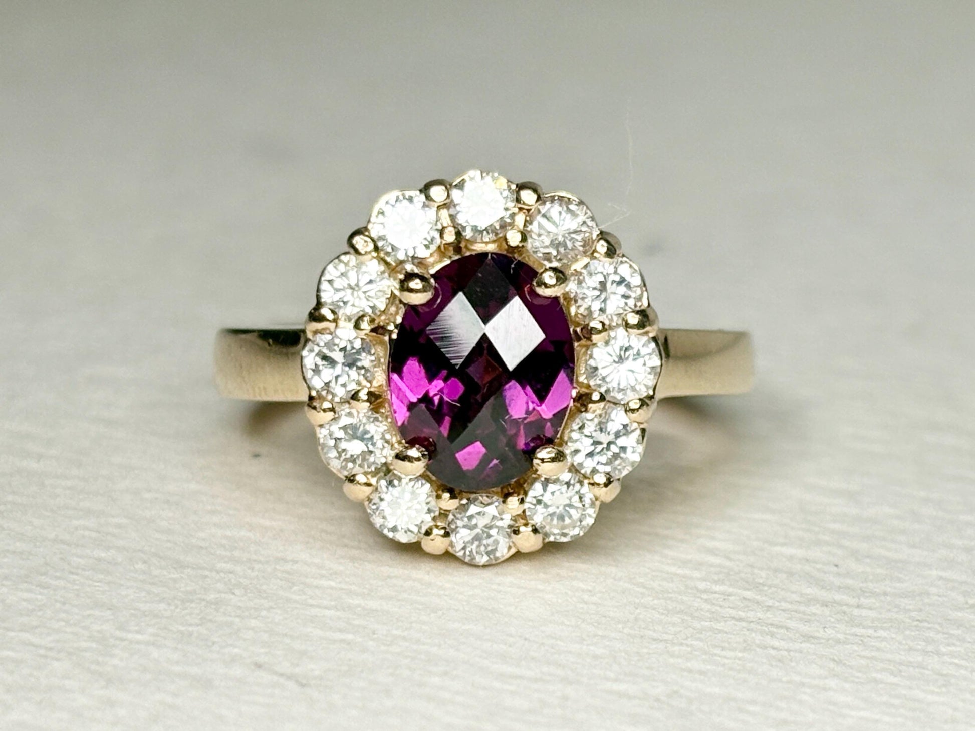 Oval checkerboard faceted rhodolite garnet ring surrounded by diamonds in a yellow gold halo setting.