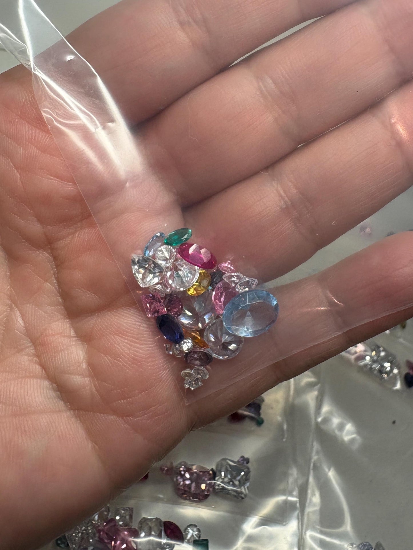 A hand holds a clear bag filled with a variety of colorful faceted gemstones.